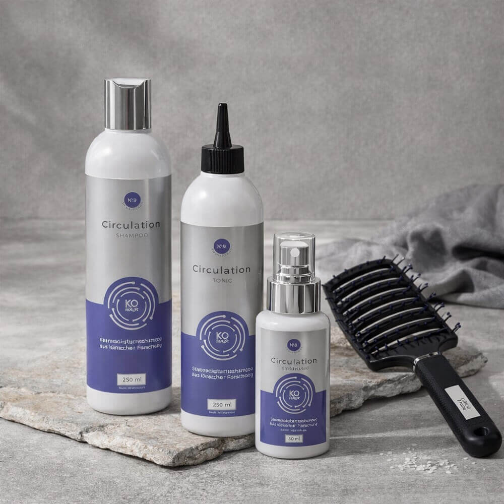 KÖ-HAIR Circulation Shampoo, Tonic und Serum Spray auf grauer Steinfläche arrangiert, daneben eine schwarze Haarbürste liegend – stilvolles, ruhiges Produkt-Setup.